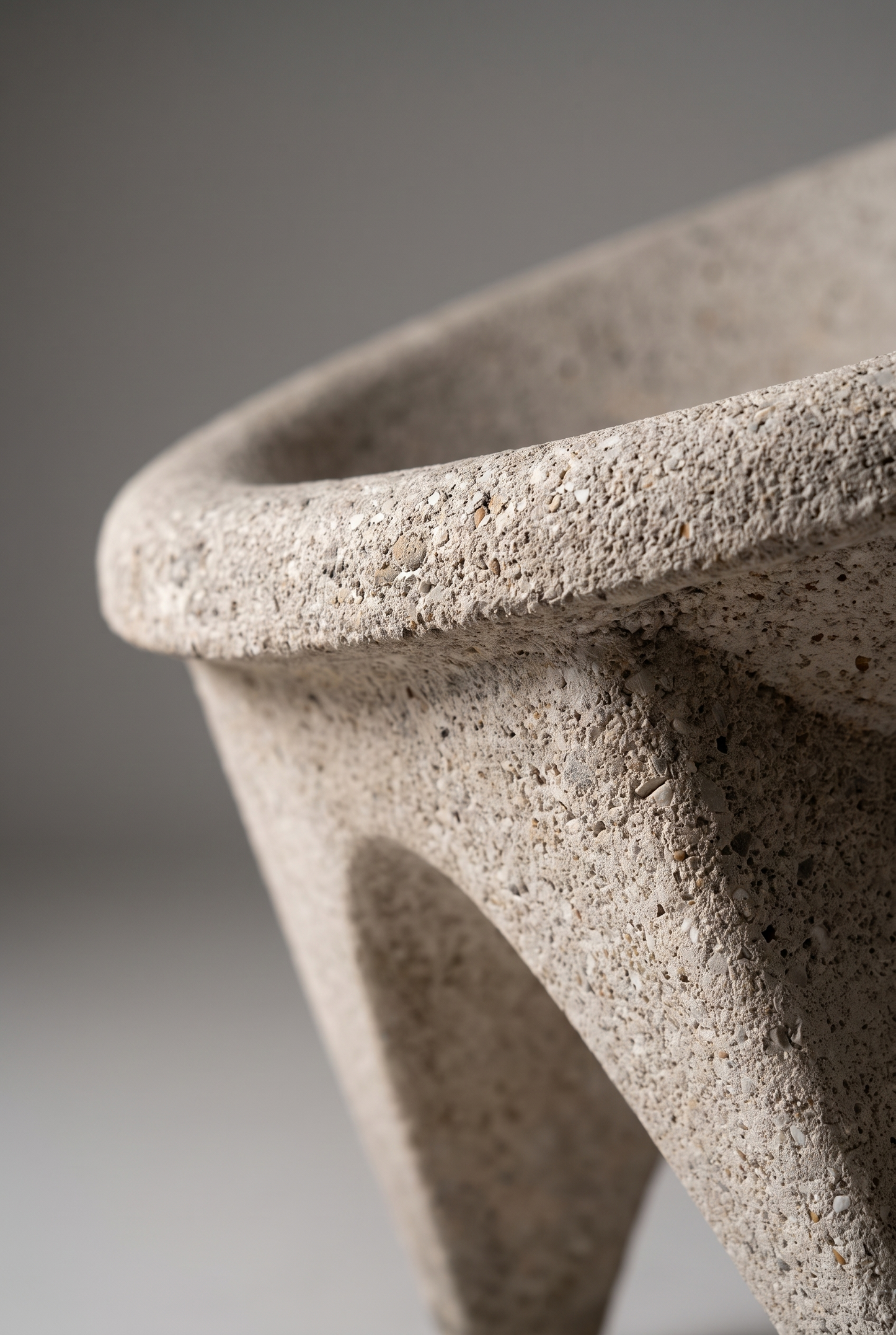 Close-up of a textured stone or concrete planter bowl with a speckled beige-gray finish, showing the curved rim and supporting leg against a neutral gray background.